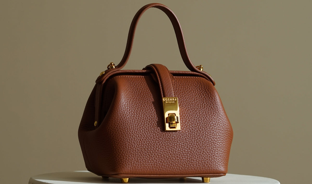 A brown BOSHRA Mahtab Mini bag pebbled leather handbag with gold-tone hardware, displayed on a round white pedestal against a warm beige background.